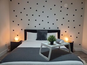 Een moderne slaapkamer met een bed, twee nachtkastjes, een bijzettafeltje, een potplant en een muur met zwarte driehoekstickers.