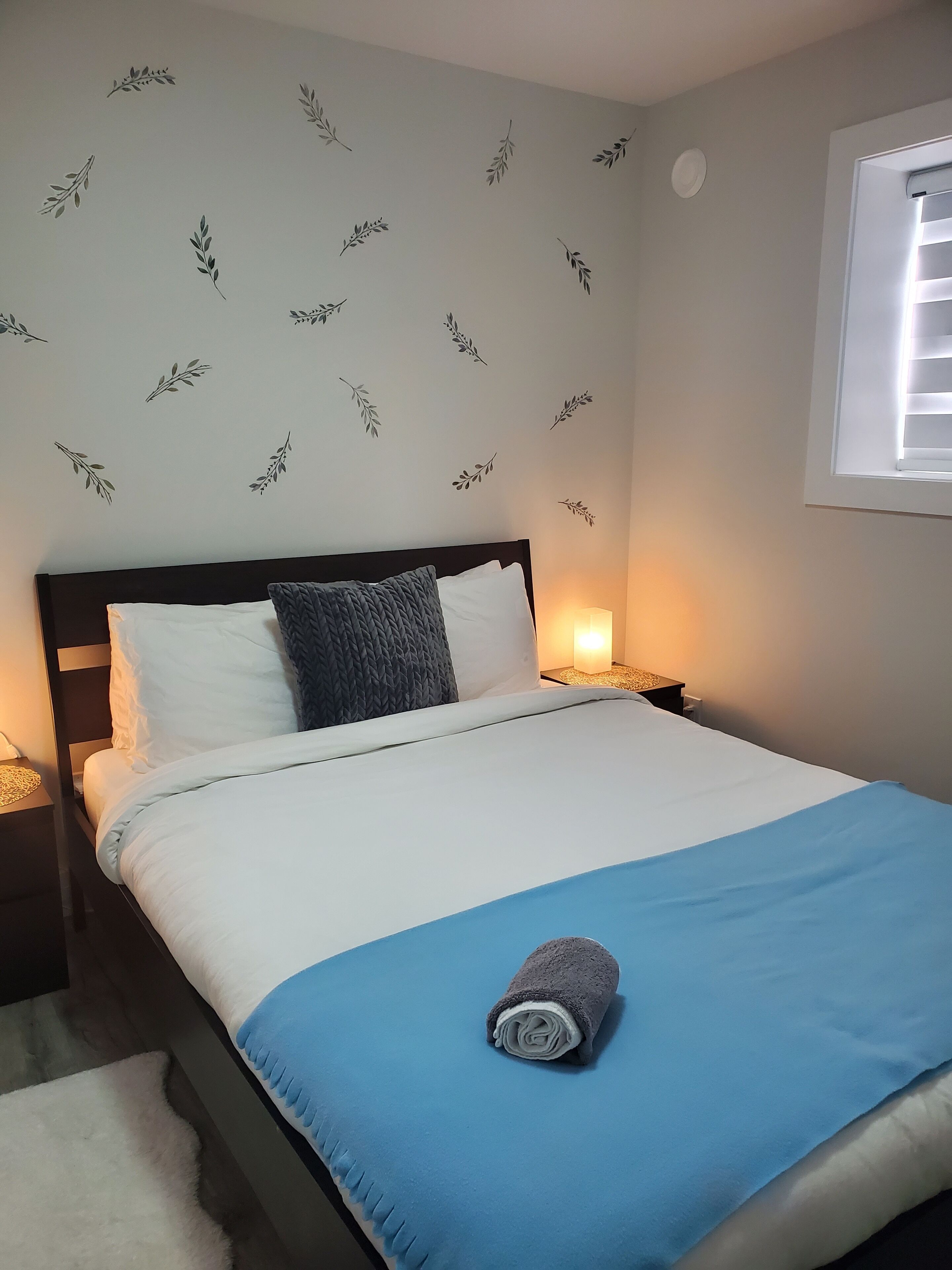 A neatly made bed with a blue bedspread and a rolled towel, a dark headboard, white pillows, and a wall with leaf patterns.