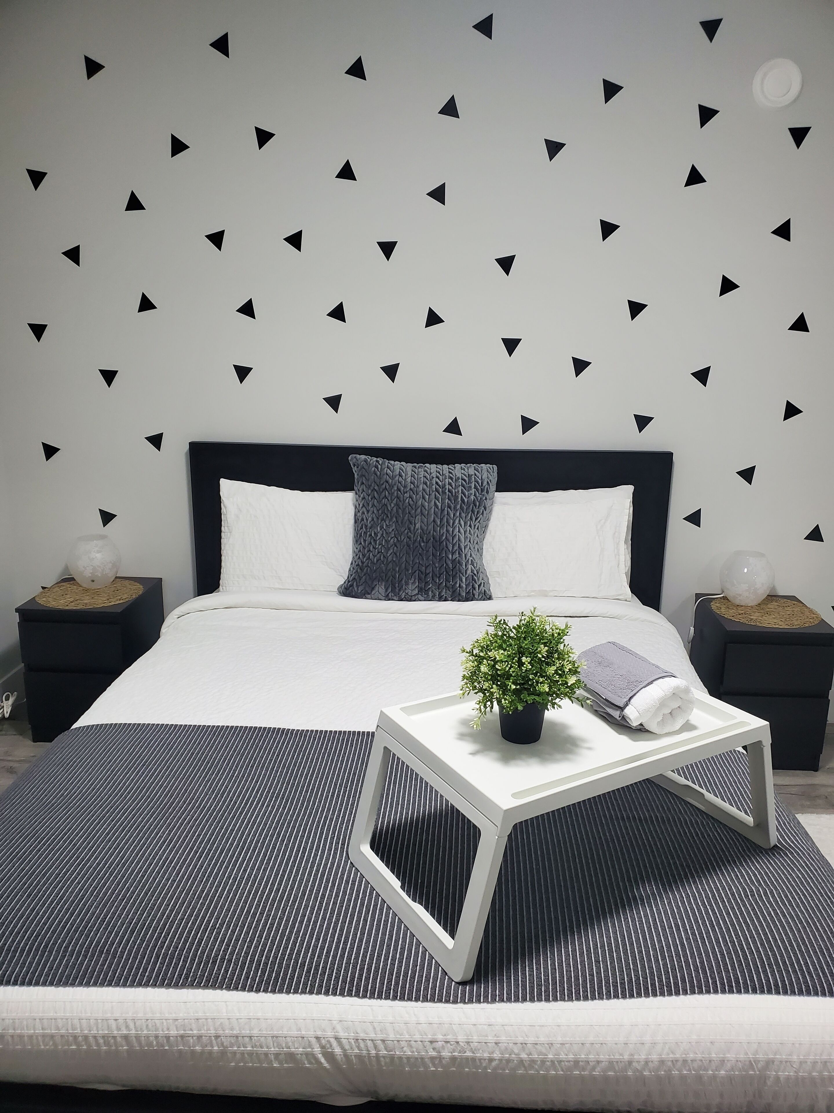 A modern bedroom with a black and white color scheme, featuring a bed with a white headboard, a grey knitted throw pillow, a small potted plant, and a white bedside table with a towel.