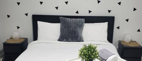 A modern bedroom with a black and white color scheme, featuring a bed with a white headboard, a grey knitted throw pillow, a small potted plant, and a white bedside table with a towel.