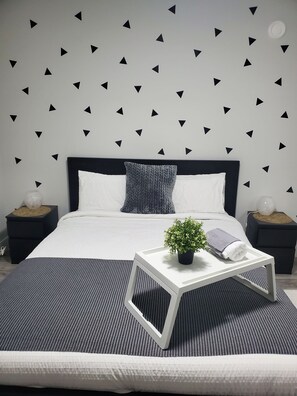 A modern bedroom with a black and white color scheme, featuring a bed with a white headboard, a grey knitted throw pillow, a small potted plant, and a white bedside table with a towel. - Lux Modern 2bd Radiant Suite+Free Parking (Delta)
