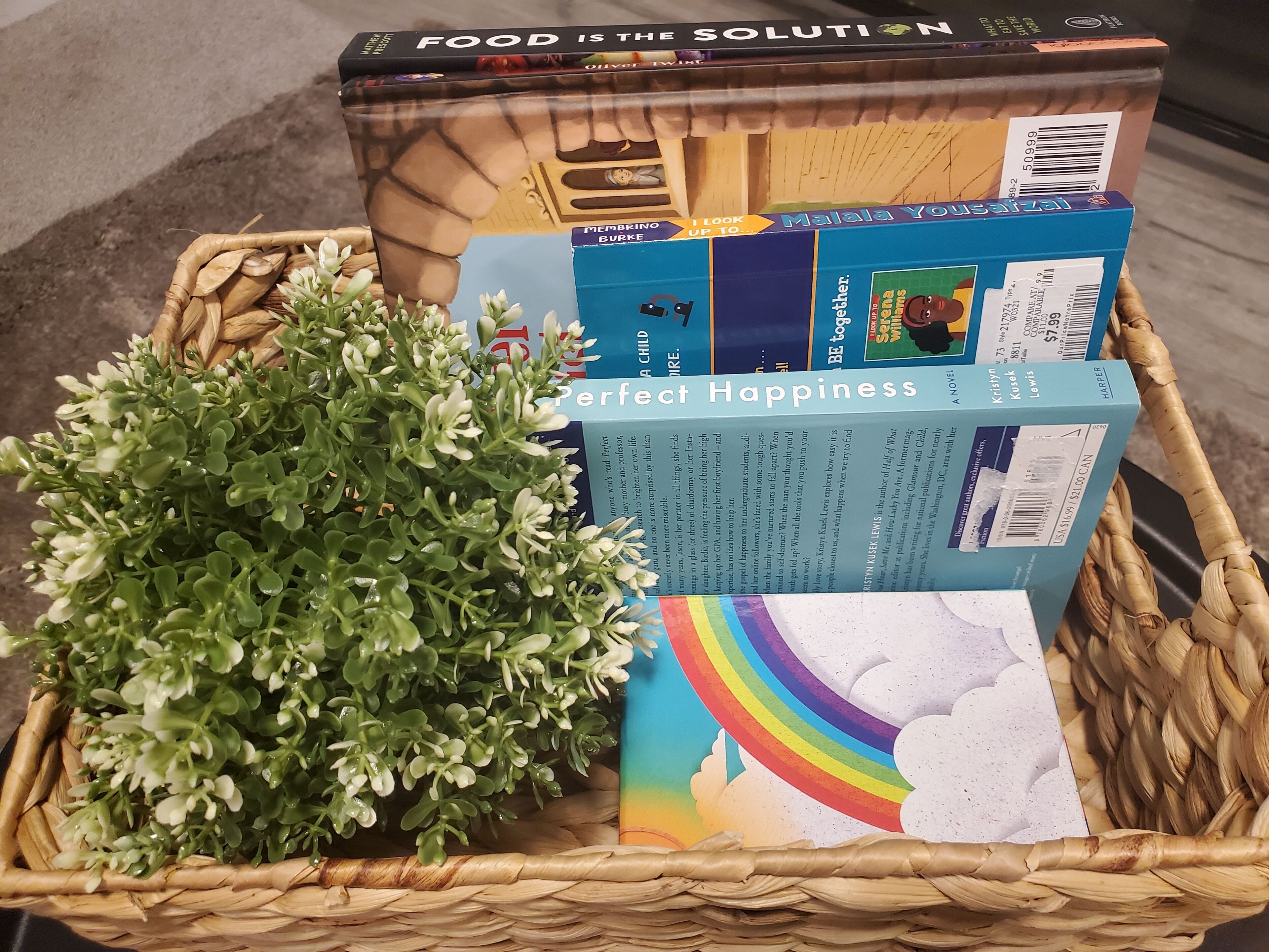 Ein Korb mit einem Buch mit dem Titel 'Perfect Happiness', einem verpackten Gegenstand mit dem Aufdruck 'Malala Yousafzai' und einer kleinen, grünen Pflanze.