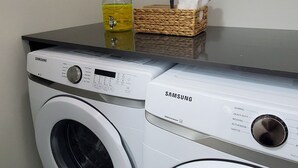 Una lavadora y secadora Samsung en un cuarto de lavado, con una botella de detergente y una cesta de toallas.