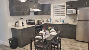 Una cocina moderna con armarios oscuros, un refrigerador de acero inoxidable, un microondas y un horno empotrado. Hay una mesa de comedor con sillas y un pequeño cubo de basura.