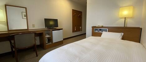 A hotel room with a bed, a desk with a television, a refrigerator, and a wardrobe.