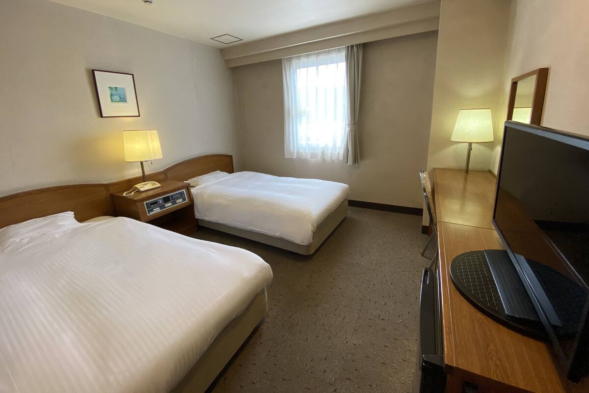 Une chambre d’hôtel avec deux lits, un bureau avec un ordinateur, une télévision, une lampe et une fenêtre avec des rideaux.