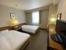 Una habitación de hotel con dos camas, un escritorio con computadora, televisor, lámpara y ventana con cortinas.