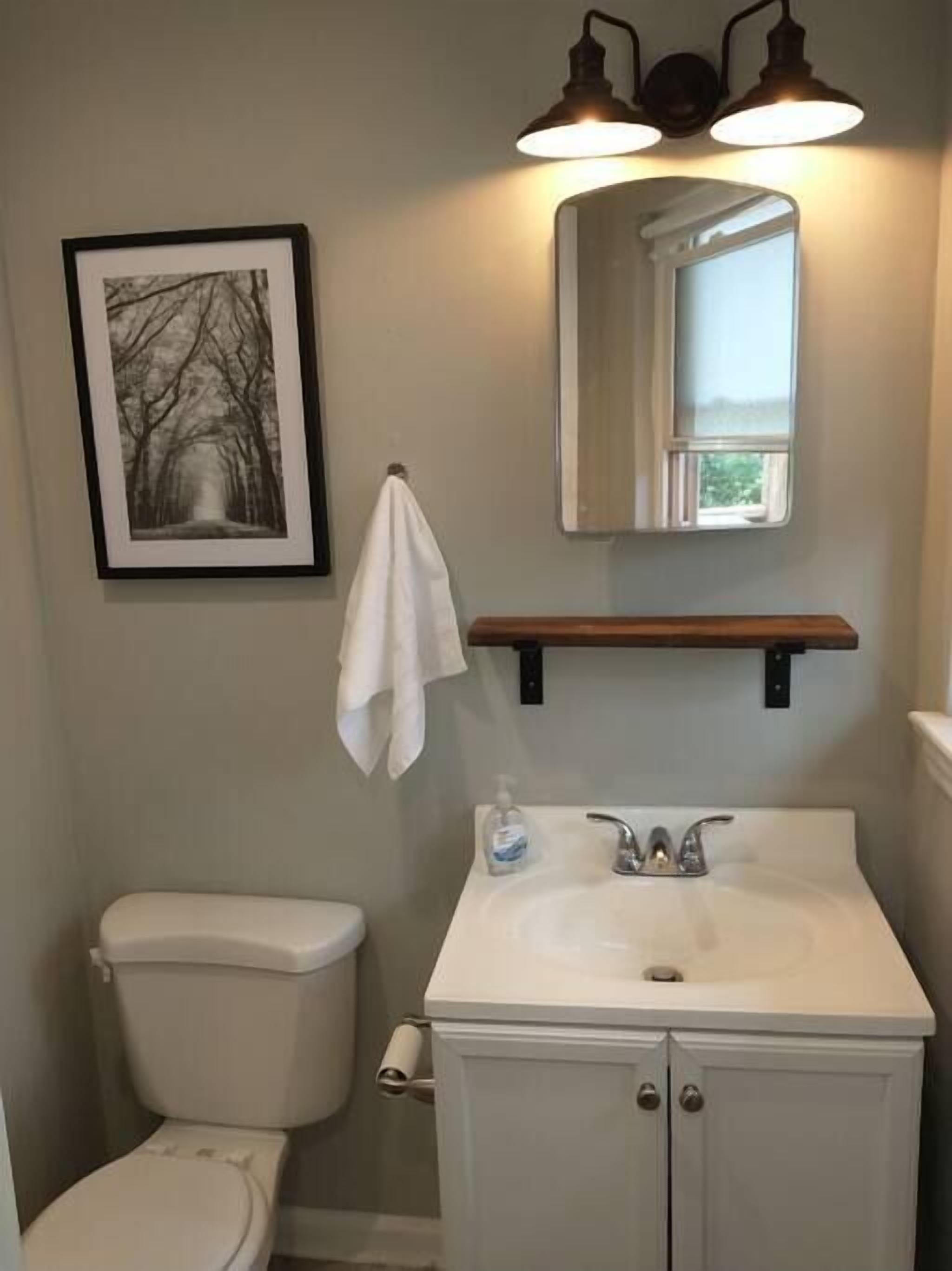 A bathroom with a toilet, sink, and mirror. There is a framed picture on the wall and a towel hanging on a hook.