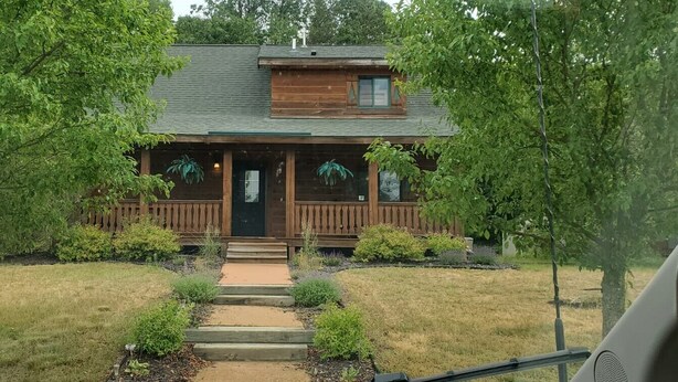 Une maison en bois à l’allure rustique, dotée d’un porche à l’avant, entourée d’arbres et d’une pelouse bien entretenue.