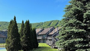 A grassy area with a wooden bench, a row of buildings with sloped roofs, and a backdrop of trees and hills.