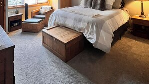 A bedroom with a large bed, two bedside tables, a window with blinds, a door, and an air conditioning unit.