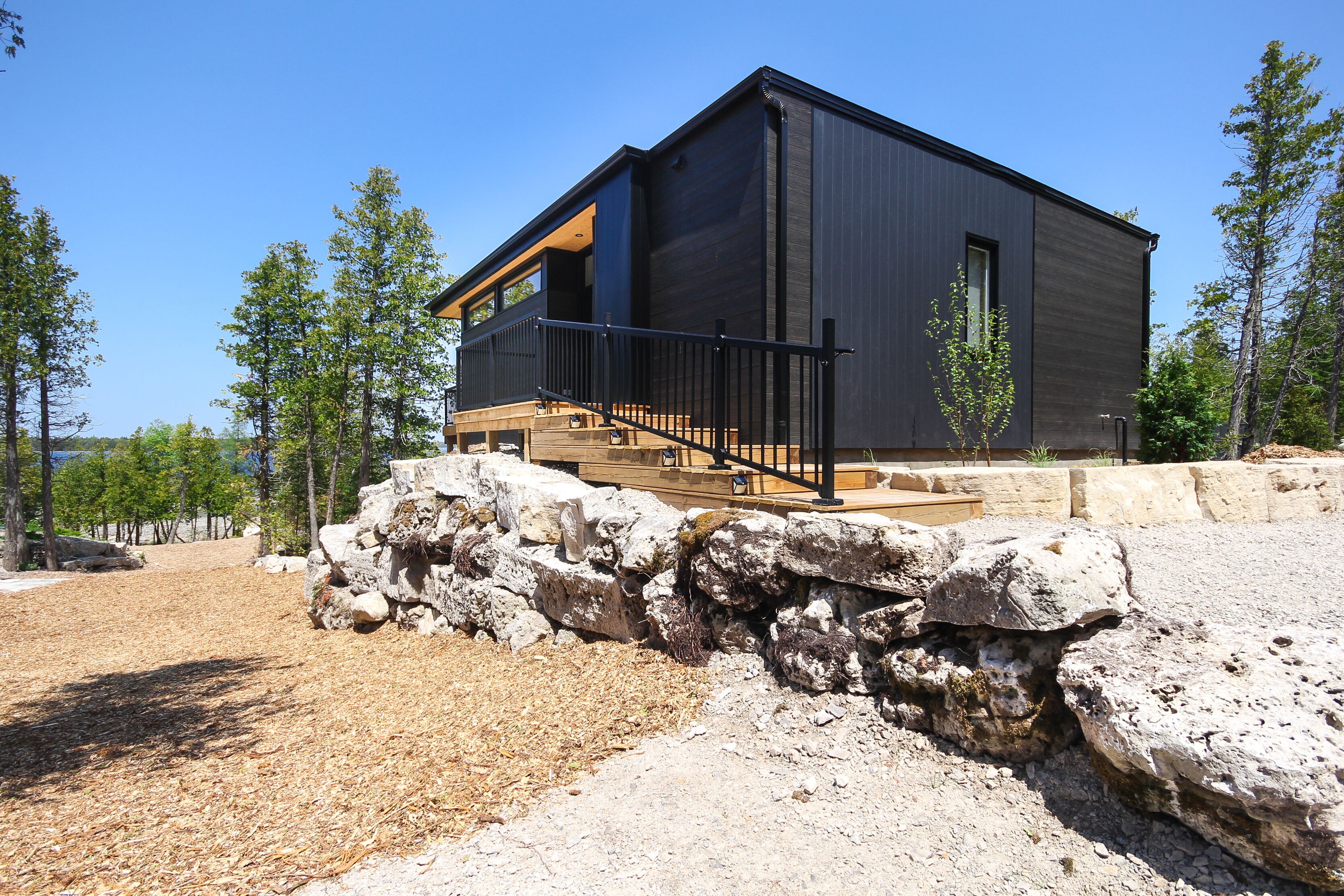 A modern two-story building with a dark exterior, a wooden deck, and a stone retaining wall.