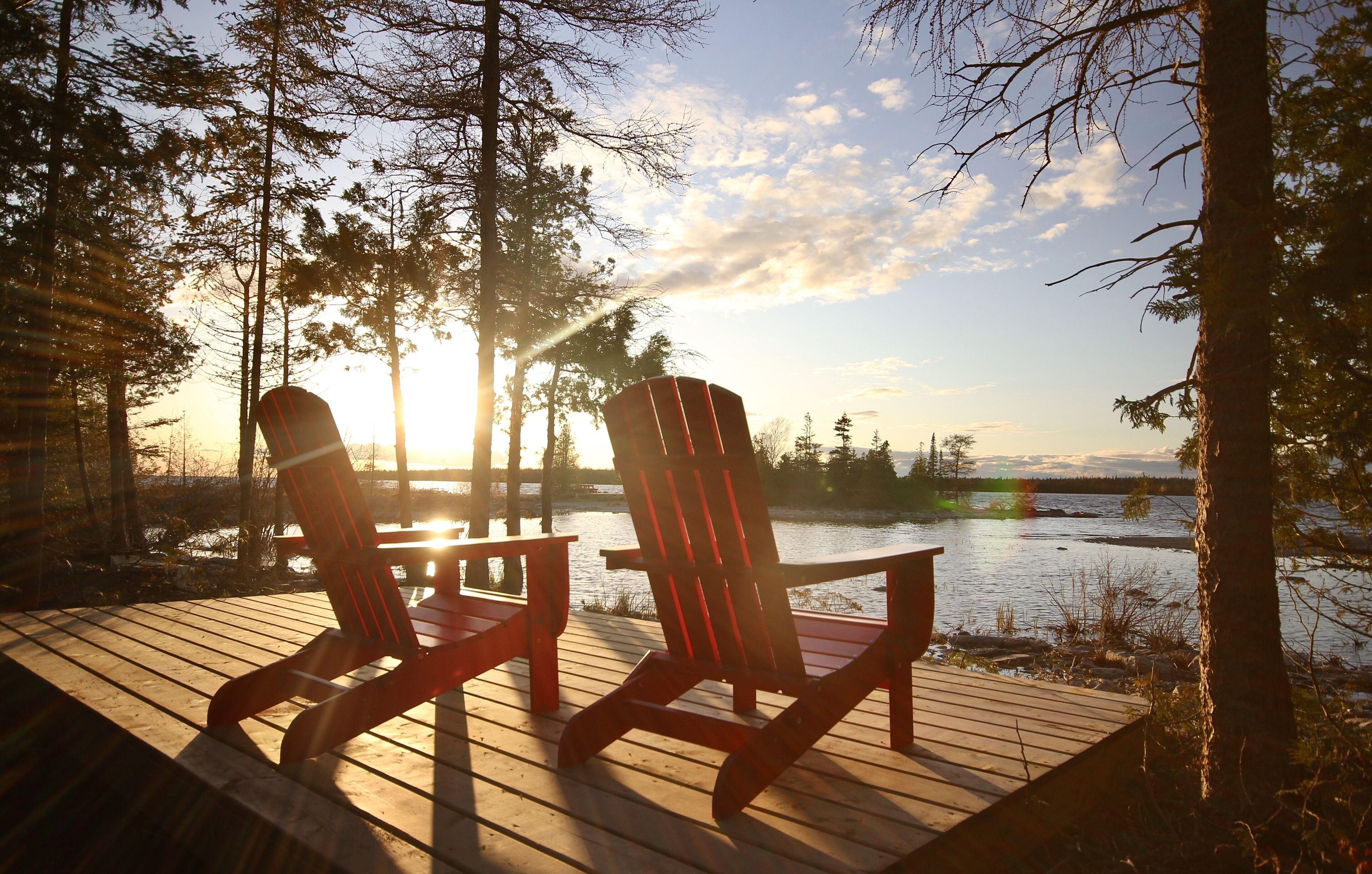 Two Adirondack chairs on a wooden deck overlooking a lake with trees and a sunset in the background.