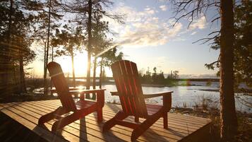 Two Adirondack chairs on a wooden deck overlooking a lake with trees and a sunset in the background.