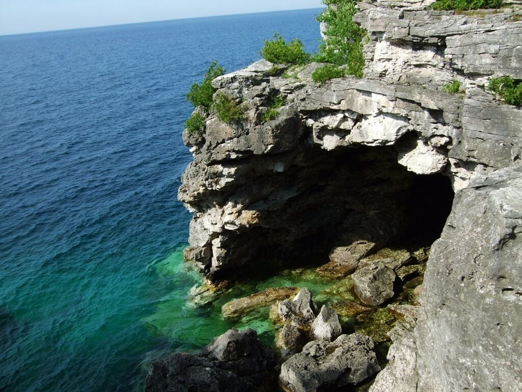 Acantilado costero rocoso con una abertura de cueva, agua cristalina y vegetación en la parte superior.