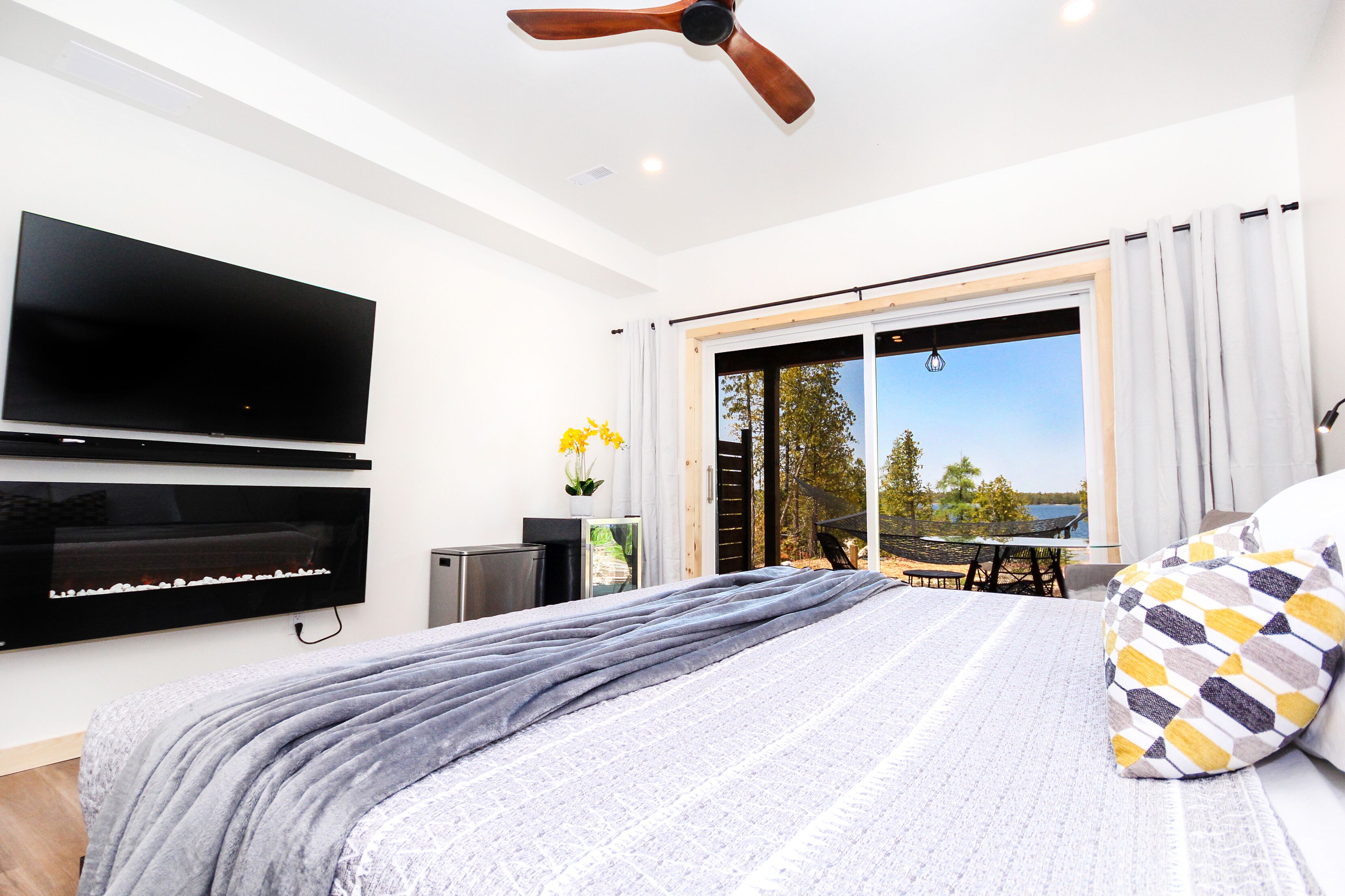 A bedroom with a bed, a flat-screen TV, a ceiling fan, a sliding glass door to a balcony, and a view of the outdoors.