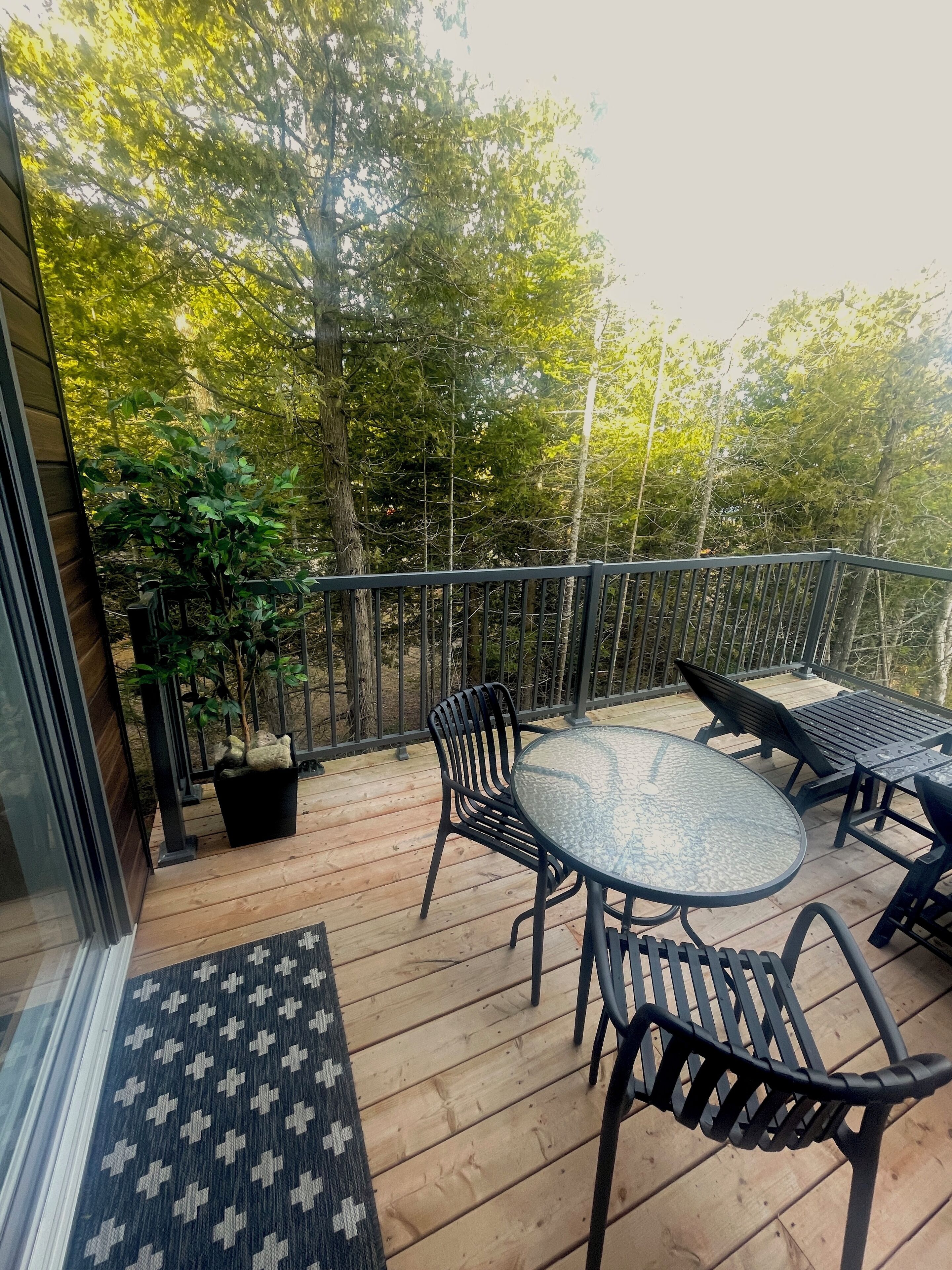 Una terraza de madera con puerta de vidrio, una mesa redonda, dos sillas y una planta en maceta.