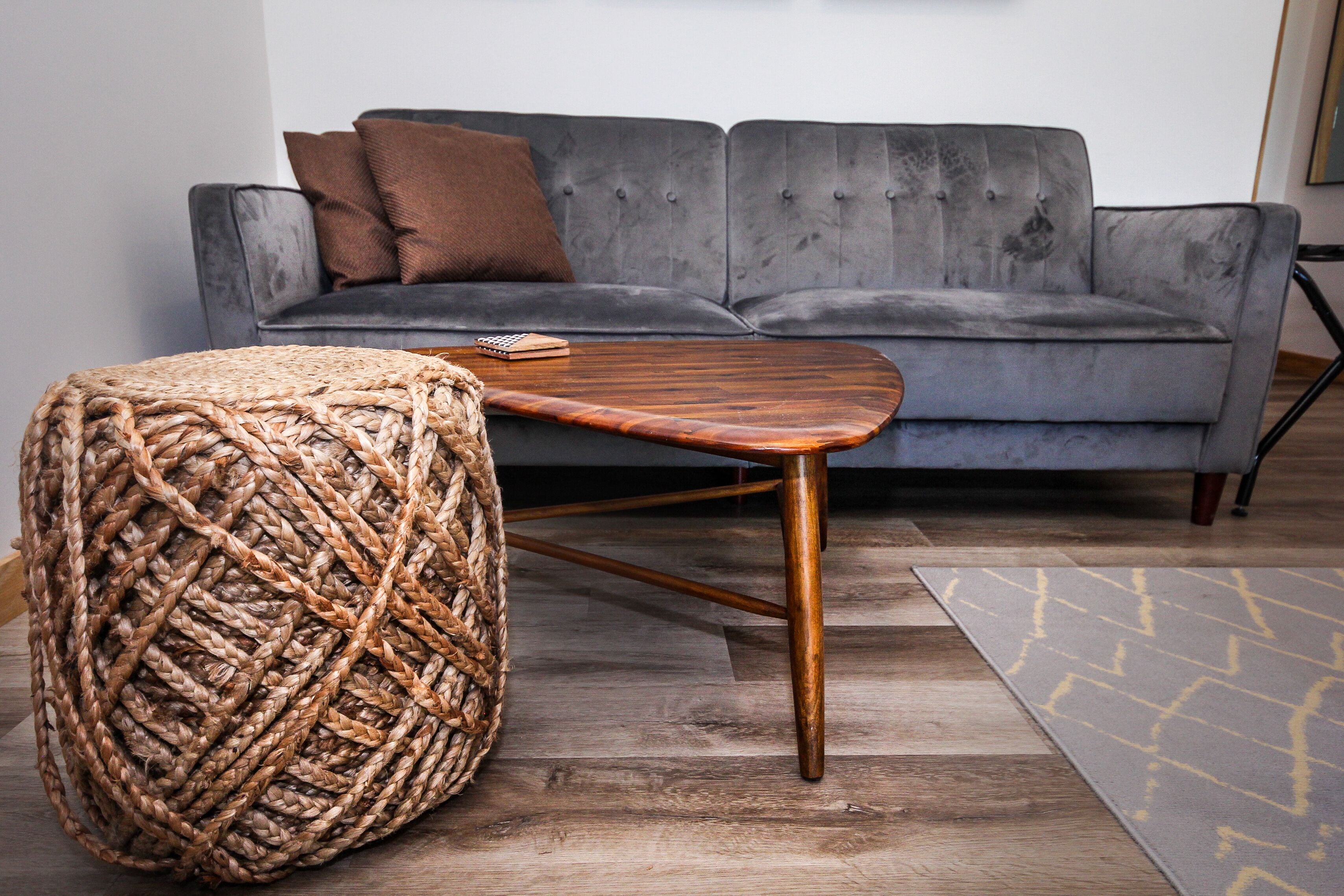 Ein graues Sofa mit einem braunen Kissen, ein gewebter Hocker und ein Couchtisch aus Holz in einem modernen Wohnzimmer mit Holzboden und einem gemusterten Teppich.