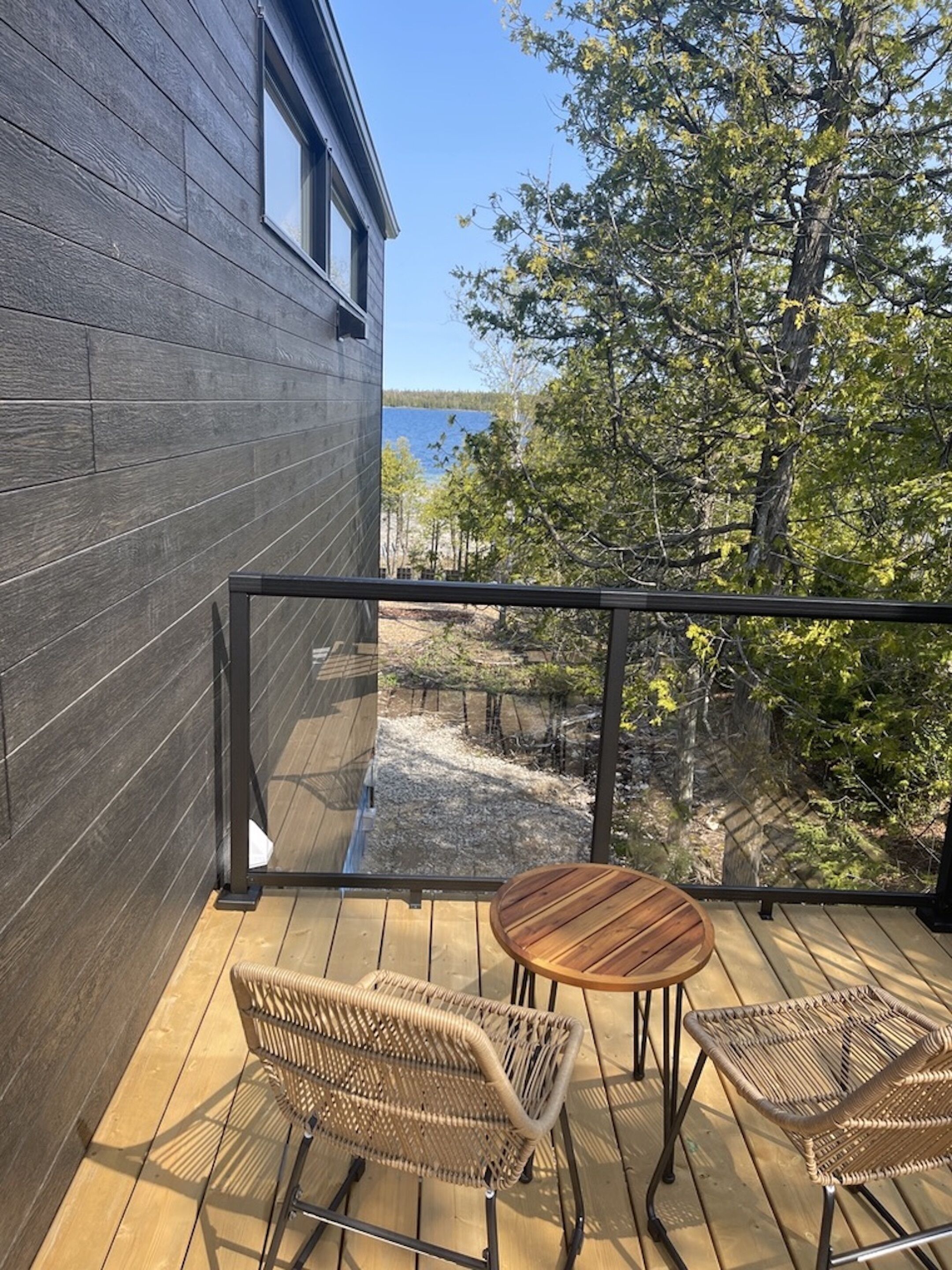 Eine Holzterrasse mit Korbmöbeln, einer Glasreling und Blick auf Bäume und ein Gewässer.