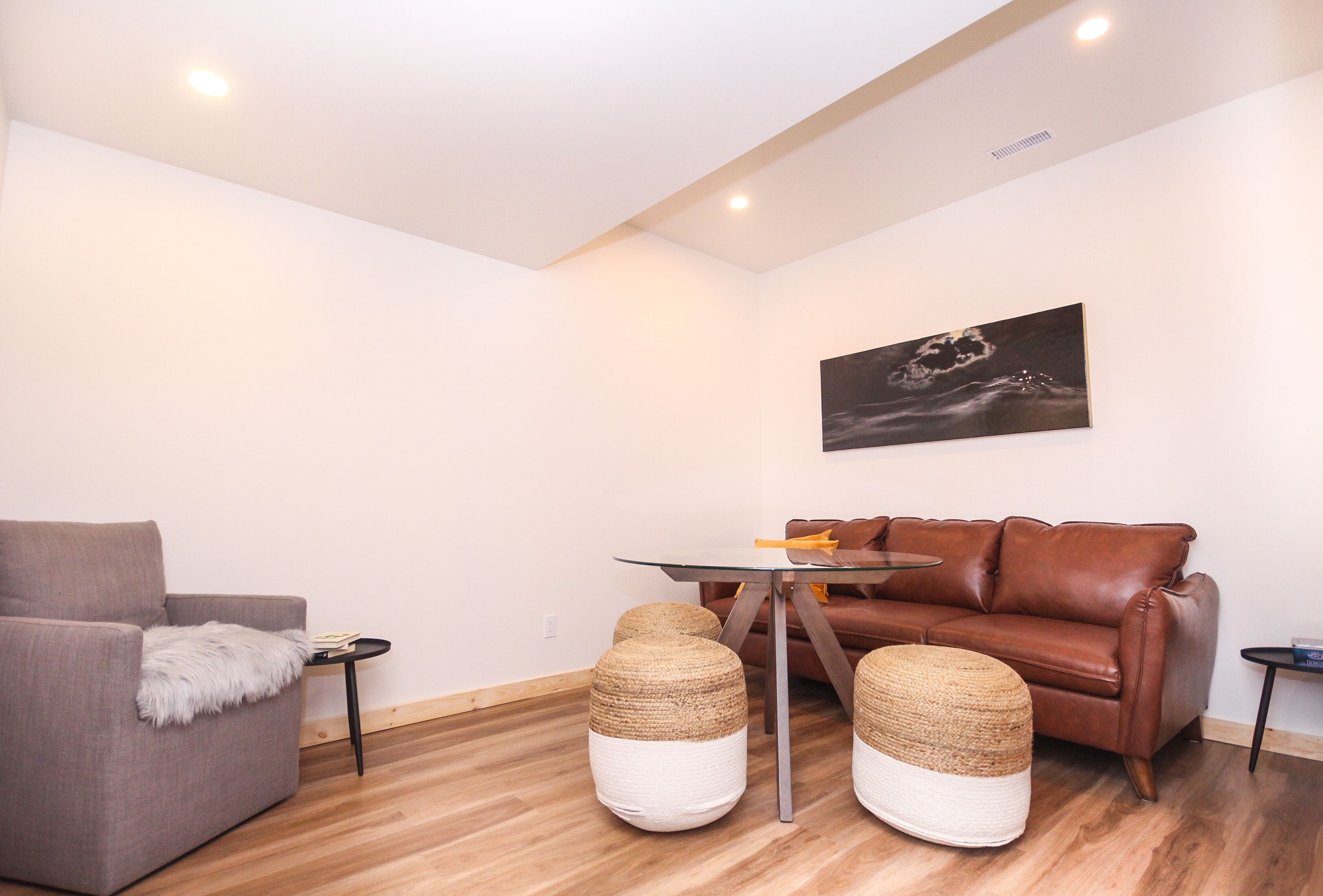 Una sala moderna con un sofá gris, un sofá de cuero marrón, una mesa de centro de vidrio y otomanas tejidas.
