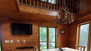 Interior de una cabaña de madera con una gran mesa de comedor, bancos, un televisor montado en la pared y un candelabro.