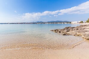 Uma praia costeira com águas cristalinas, costa rochosa e edifícios ao longe.