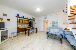 Uma sala de estar com sofá, mesa de jantar com cadeiras, televisão e área de cozinha com armários e geladeira.