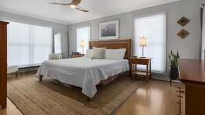 A spacious bedroom with a large bed, two bedside tables with lamps, a ceiling fan, and a view of the outdoors through large windows. - Landhaus Eichenbaum | Pool | Hot Tub (Fredericksburg)