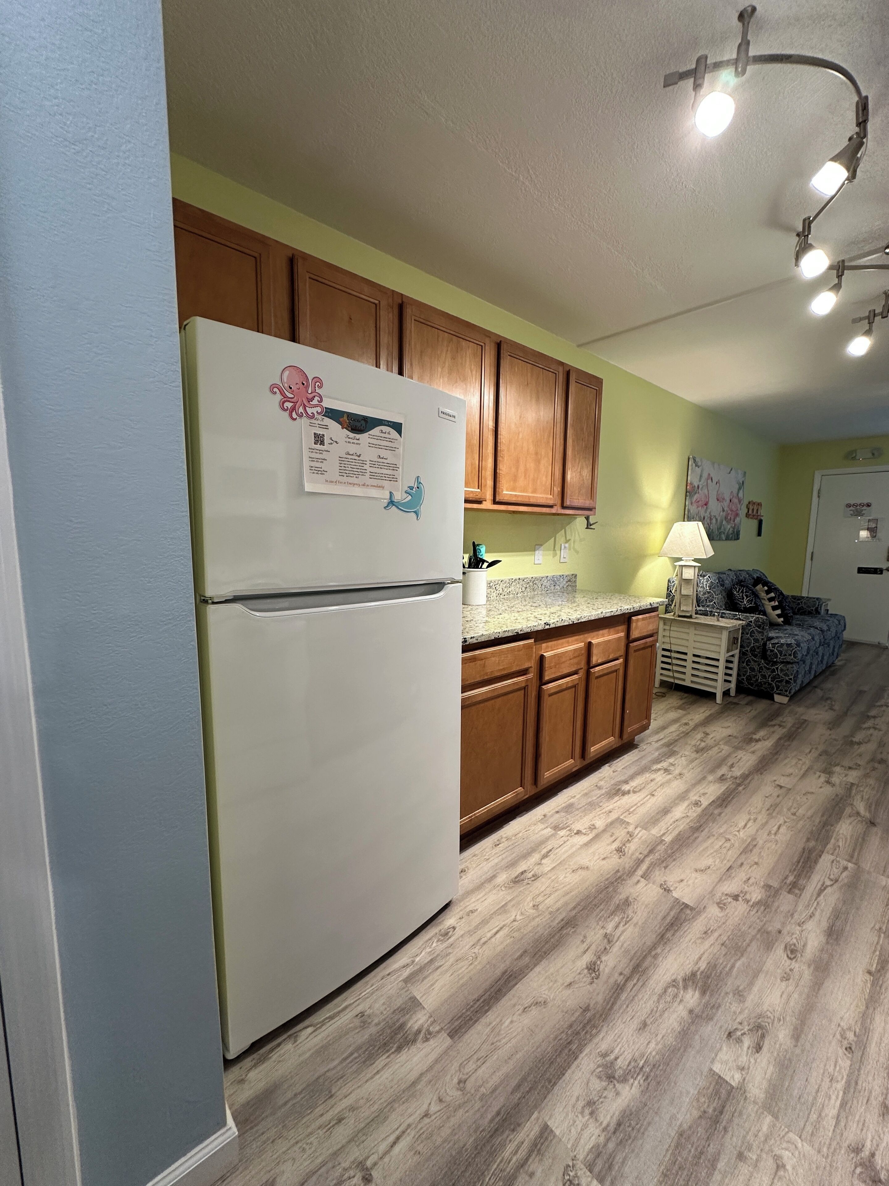 A kitchen with wooden cabinets, a white refrigerator, a granite countertop, and a dining area with a patterned sofa.