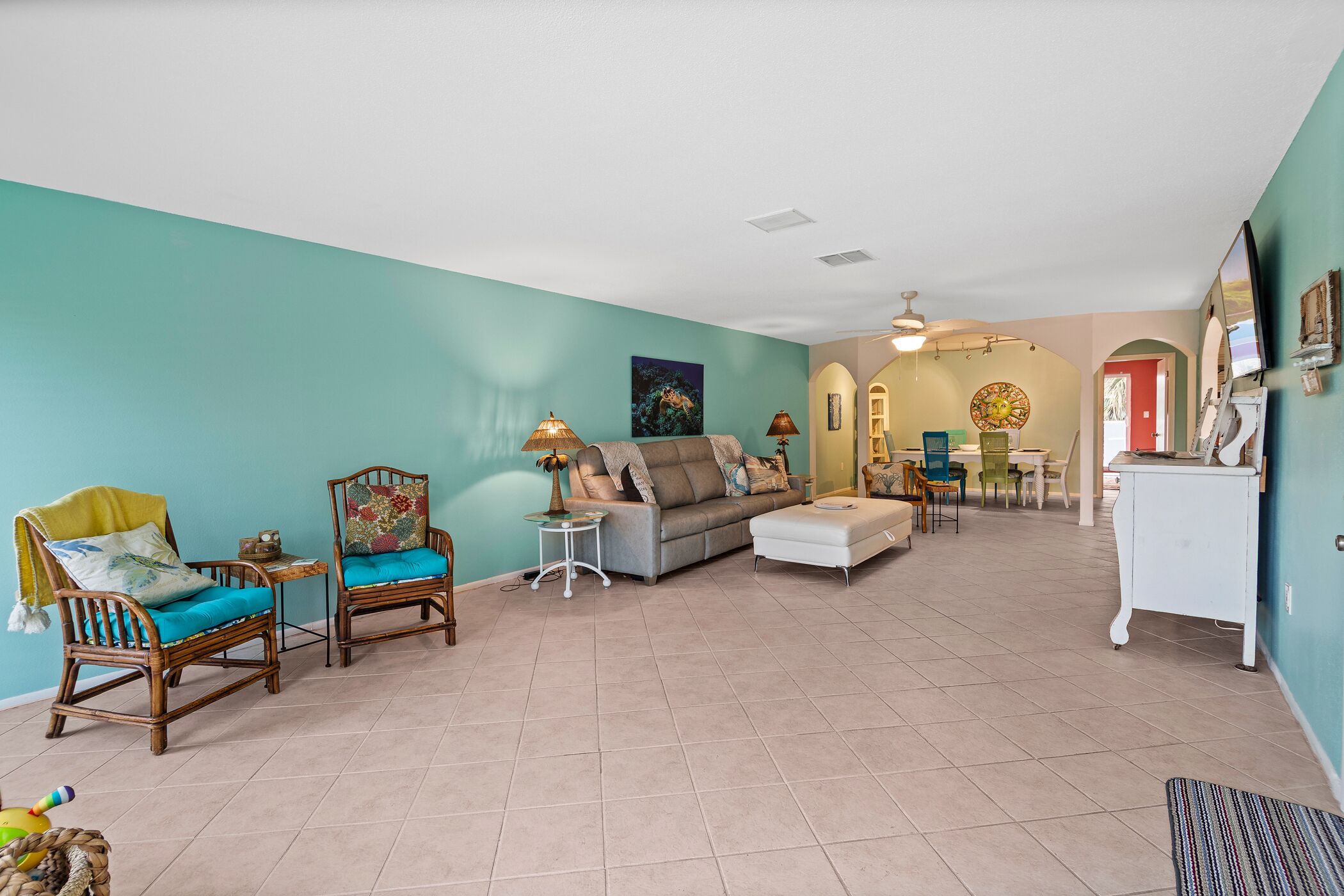 A spacious living room with teal walls, a grey sofa, two wooden chairs, and a white ottoman. There is a dining area in the background with a table and chairs.