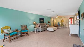 A spacious living room with teal walls, a grey sofa, two wooden chairs, and a white ottoman. There is a dining area in the background with a table and chairs.