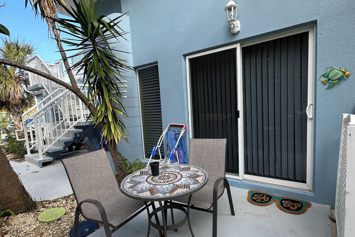 A patio with a round table and chairs, a covered area, and a building with a blue exterior and a large window.
