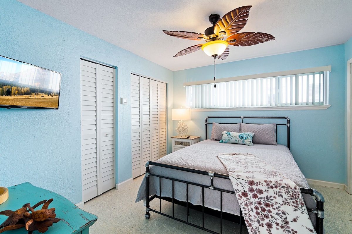 A bedroom with a bed, a ceiling fan, a window with blinds, a bedside table with a lamp, and a wall-mounted TV.