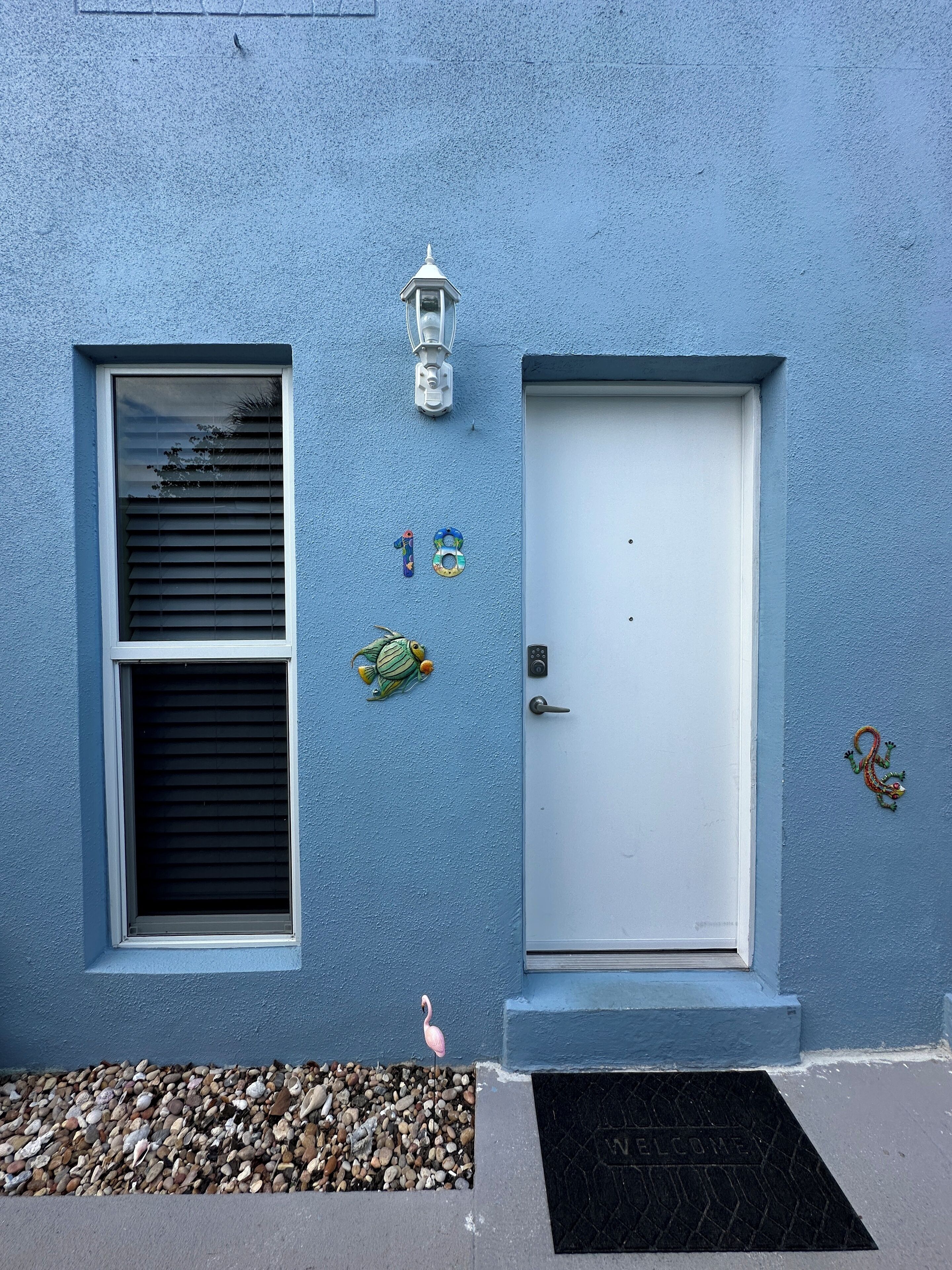 Una casa azul con puerta blanca, una ventana con persianas, un pequeño porche con guijarros y una decoración de flamenco.