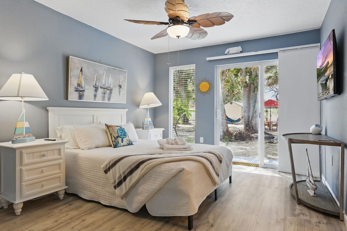 A bedroom with a bed, nightstands, a ceiling fan, and a view of the outdoors.