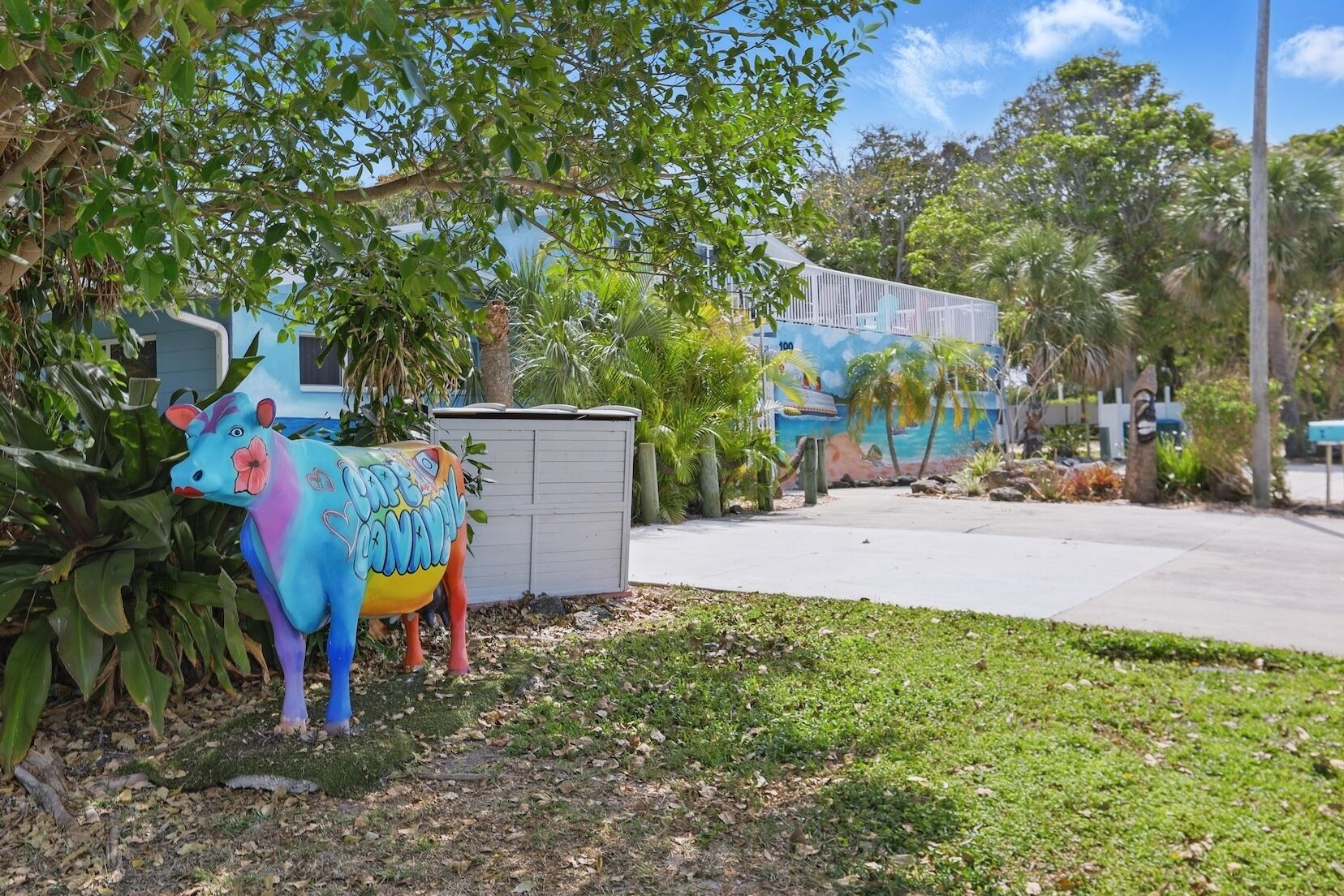 Une sculpture colorée représentant une vache, située dans un jardin, avec une fresque murale en arrière-plan.