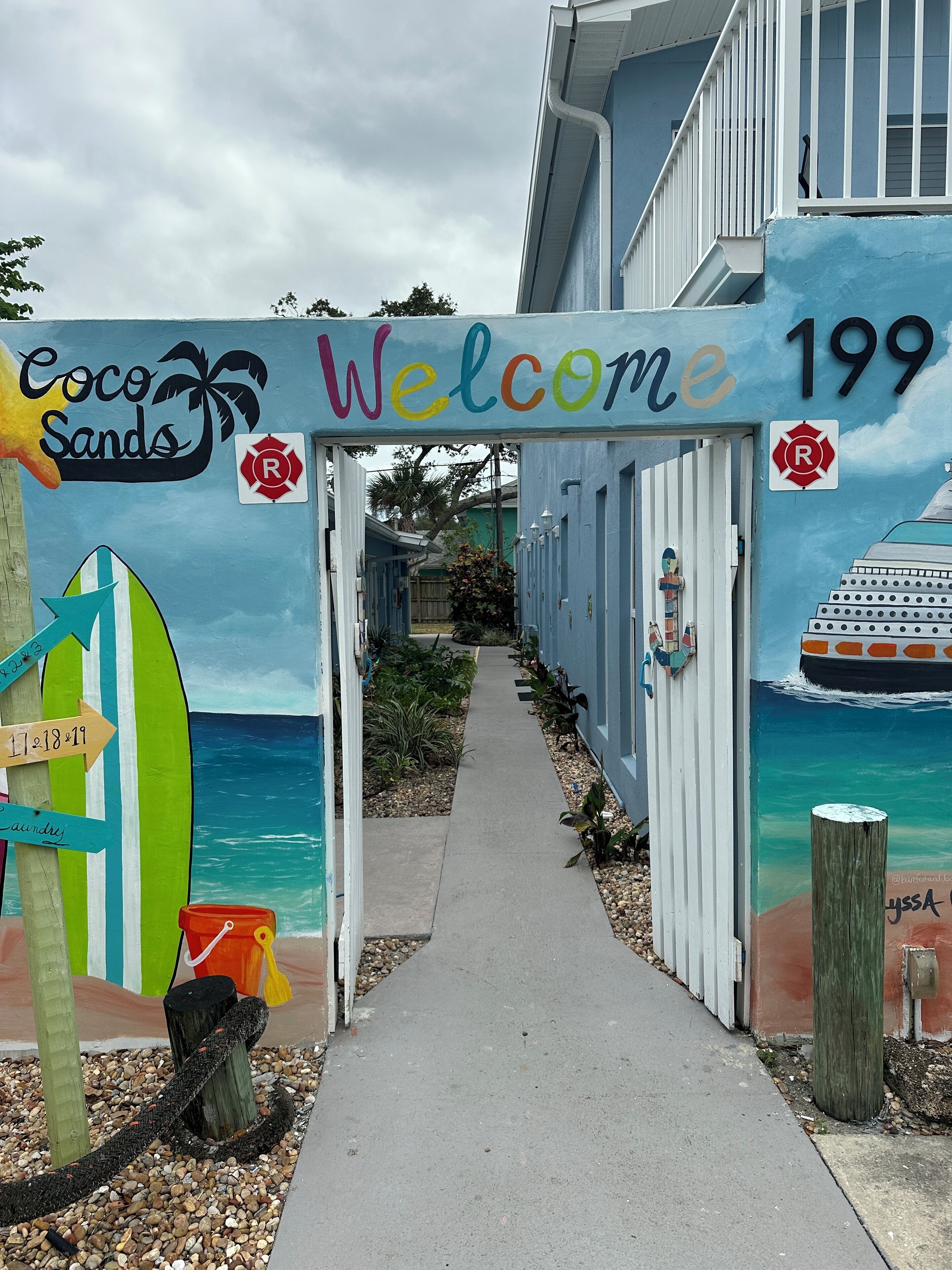 A colorful entrance with a 'Welcome' sign, a surfboard sign, and a beach-themed mural.