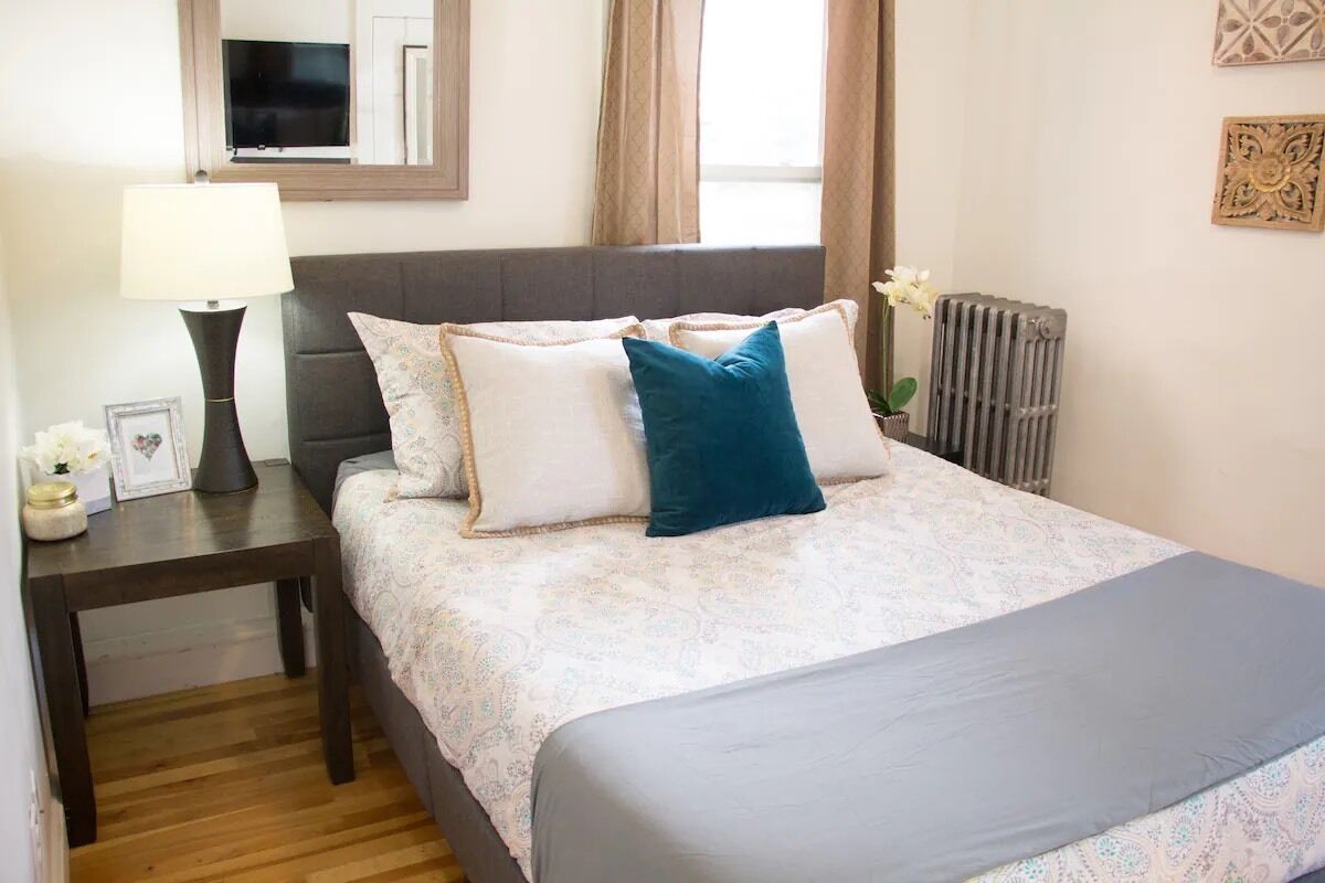 A neatly made bed with patterned bedding, a bedside table with a lamp, and a radiator.