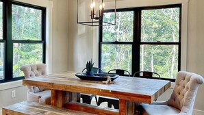 Een eetruimte met een houten tafel en banken, een geometrische hanglamp, en grote ramen met uitzicht op bomen.