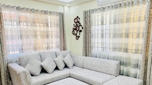 Ein Wohnzimmer mit grauem Ecksofa, passendem Hocker und gemusterten Vorhängen.