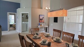 Un coin repas avec une table en bois dressée pour quatre personnes, une cuisine en arrière-plan et une grande fenêtre avec des stores.