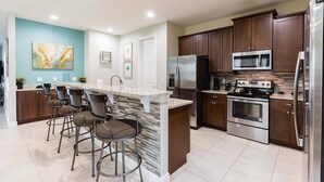 A modern kitchen with a central island, bar stools, stainless steel appliances, and a teal accent wall. - Cheery Villa with Games Room at Encore (Kissimmee)
