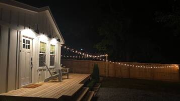 Una terraza de madera con un banco y una pequeña mesa, un edificio blanco con una puerta y ventanas, y una cerca de madera con luces colgantes.