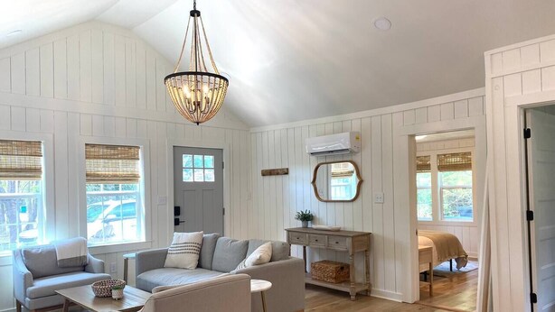 A spacious living room with a high ceiling, wooden floors, and a chandelier. There are two sofas, a coffee table, and a small side table. A wooden cabinet and a mirror are also visible. - The Belmont Cottage - 2 bedroom (Belmont)