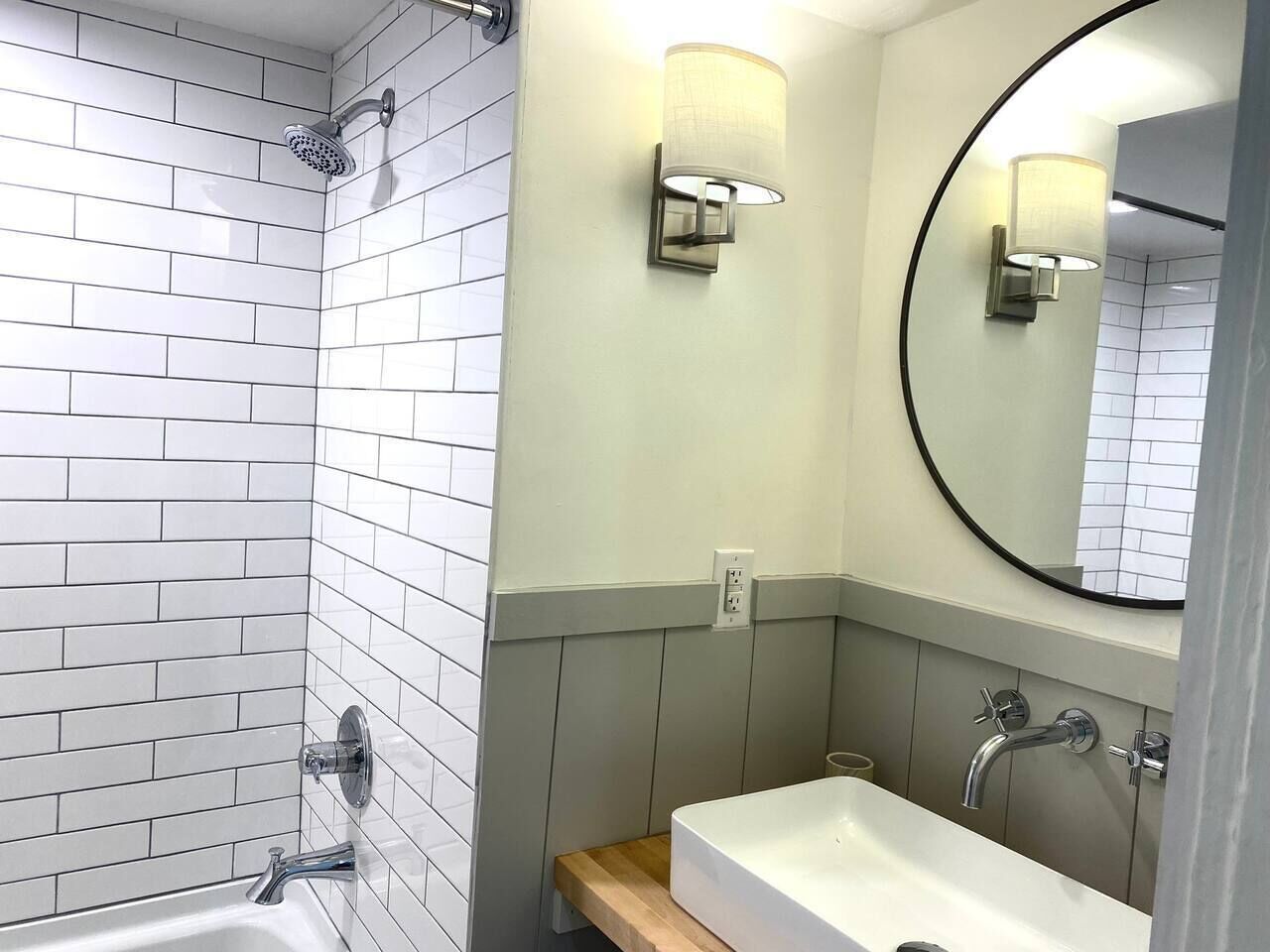 Une salle de bain moderne avec des carreaux blancs et gris, une douche, un lavabo avec un miroir et des luminaires fixés au mur.