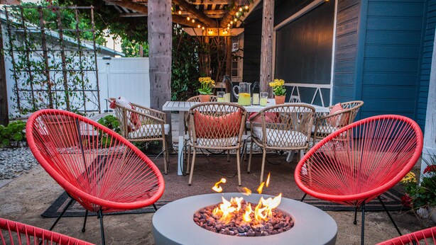 Een terras buiten met een ronde vuurkorf, rieten stoelen en een eettafel.