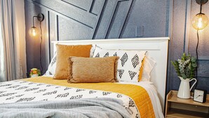 Een net opgemaakt bed met patroongedekte lakens, een nachtkastje met een klok en een wandlamp.