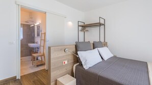 A bedroom with a bed, a wooden headboard, a bedside table, a hanging shelf, and a bathroom visible through an open door.