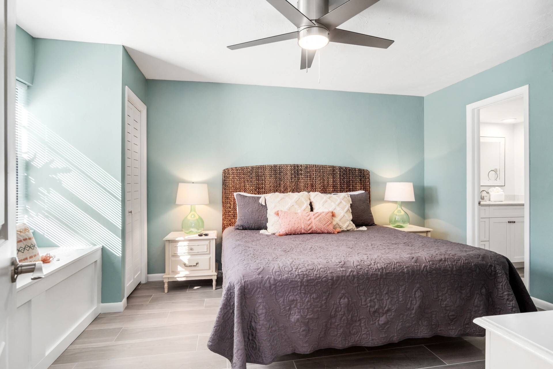 Une chambre à coucher comprenant un lit, deux tables de chevet, un ventilateur de plafond et une fenêtre avec des stores.