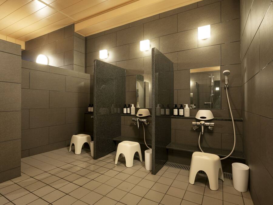 A modern bathroom with dark tiled walls, white stools, a mirror, and a shower area.